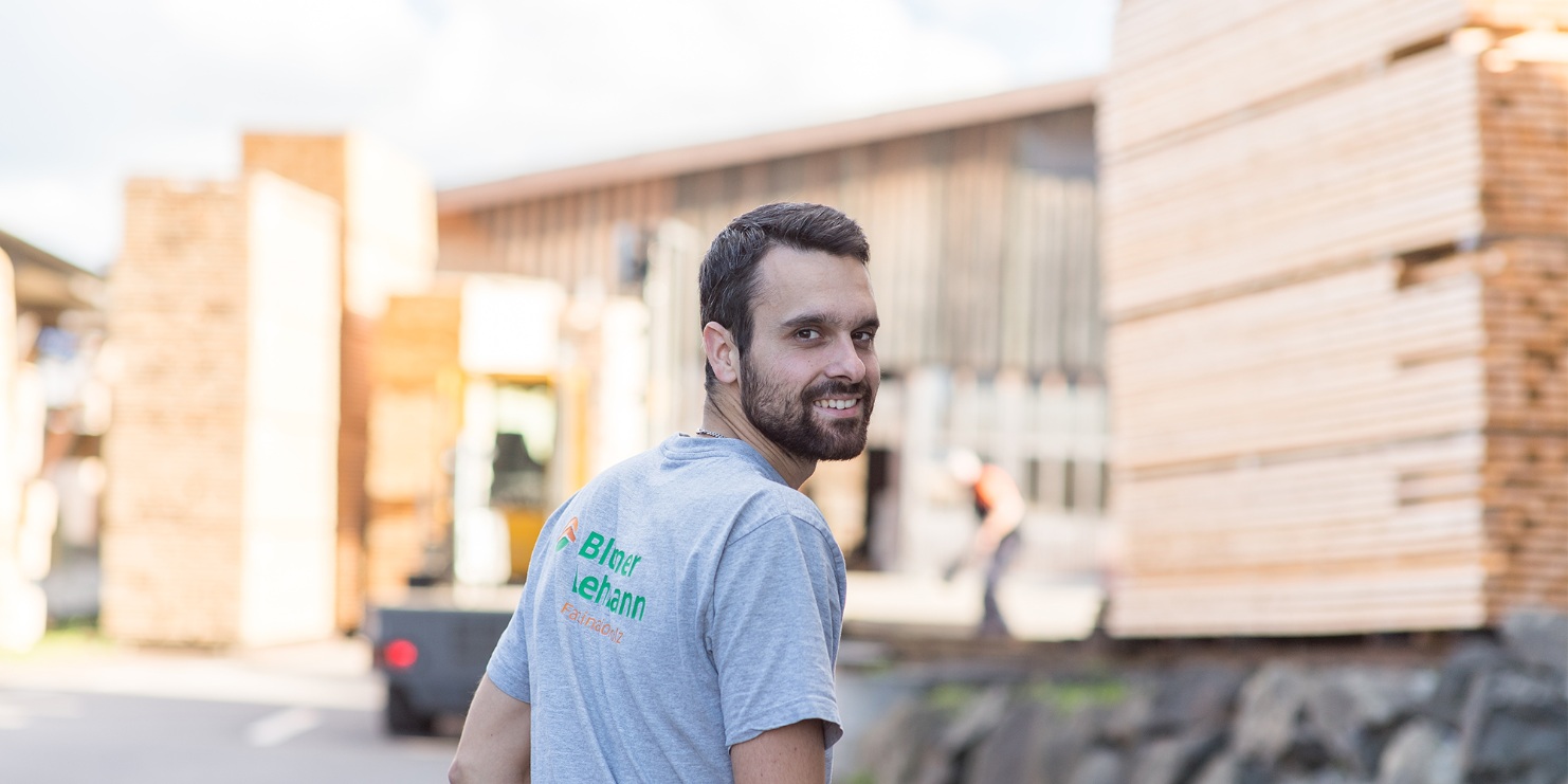 A young employee with dark hair and a beard and wearing a grey T-shirt, looking over his shoulder at the camera. A pile of cut wood can be seen in the background.