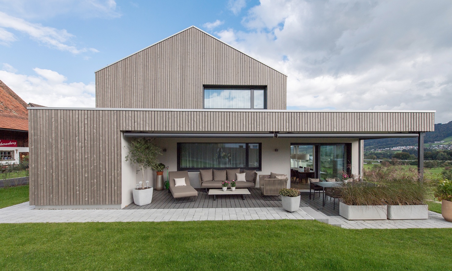 The photograph shows the single-family house with lake views and the covered seating area in the garden in fine weather