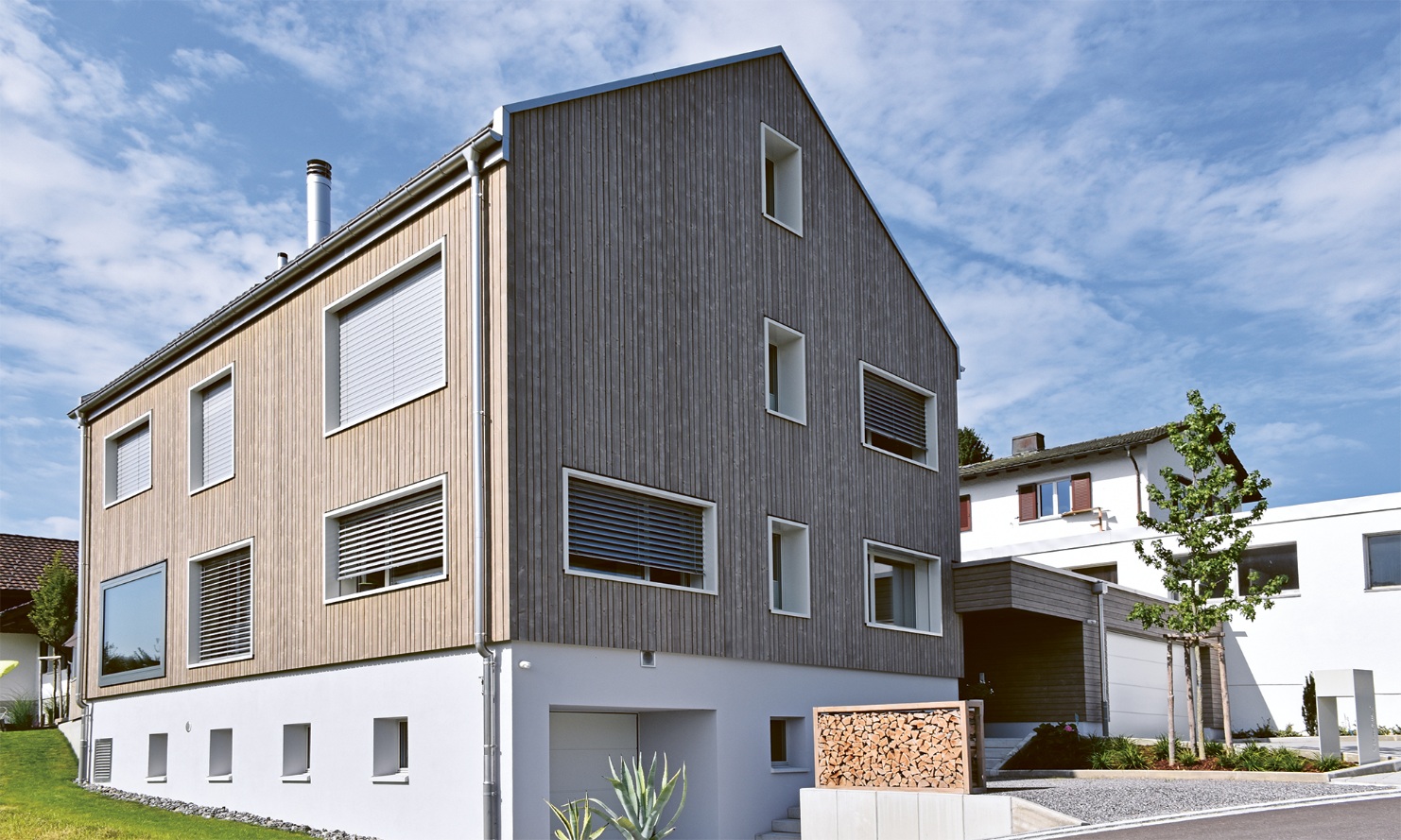 Gesamtaufnahme vom lichtdurchfluteten Einfamilienhaus mit Garten und einer Raumgestalt gefüllt mit Holzscheiter auf dem Kiesvorplatz