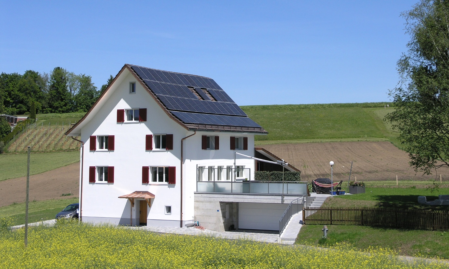 Frontaufnahme vom Einfamilienhaus mit einer umfassenden Sanierung inmitten einer begrünten Umgebung sowie einer weiten Blumenwiese vor dem Eingangsbereich bei schönem Wetter