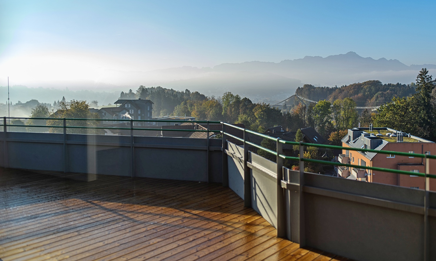 Aufnahme auf der Dachterrasse mit Holzboden des Mehrfamilienhauses Berg mit einer spektakulären Aussicht über die Hausdächer bis hin zum Bergpanorama