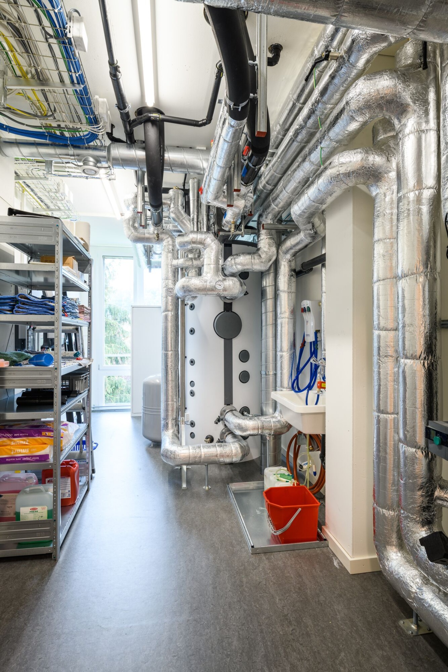 Innenansicht des Technikraumes im Schulhaus Grenzhof Luzern mit Rohren, Leitungen und Regalen