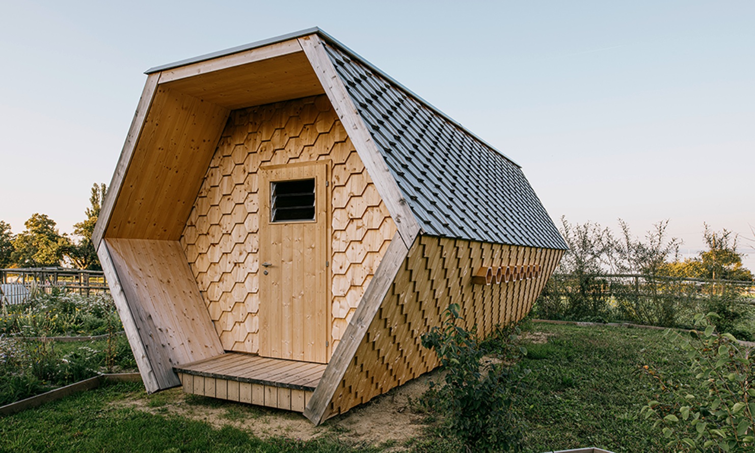 Wabenförmiges Bienenhaus mit Fassade aus sechseckigen Holzschindeln inmitten einer blühenden Wiese.