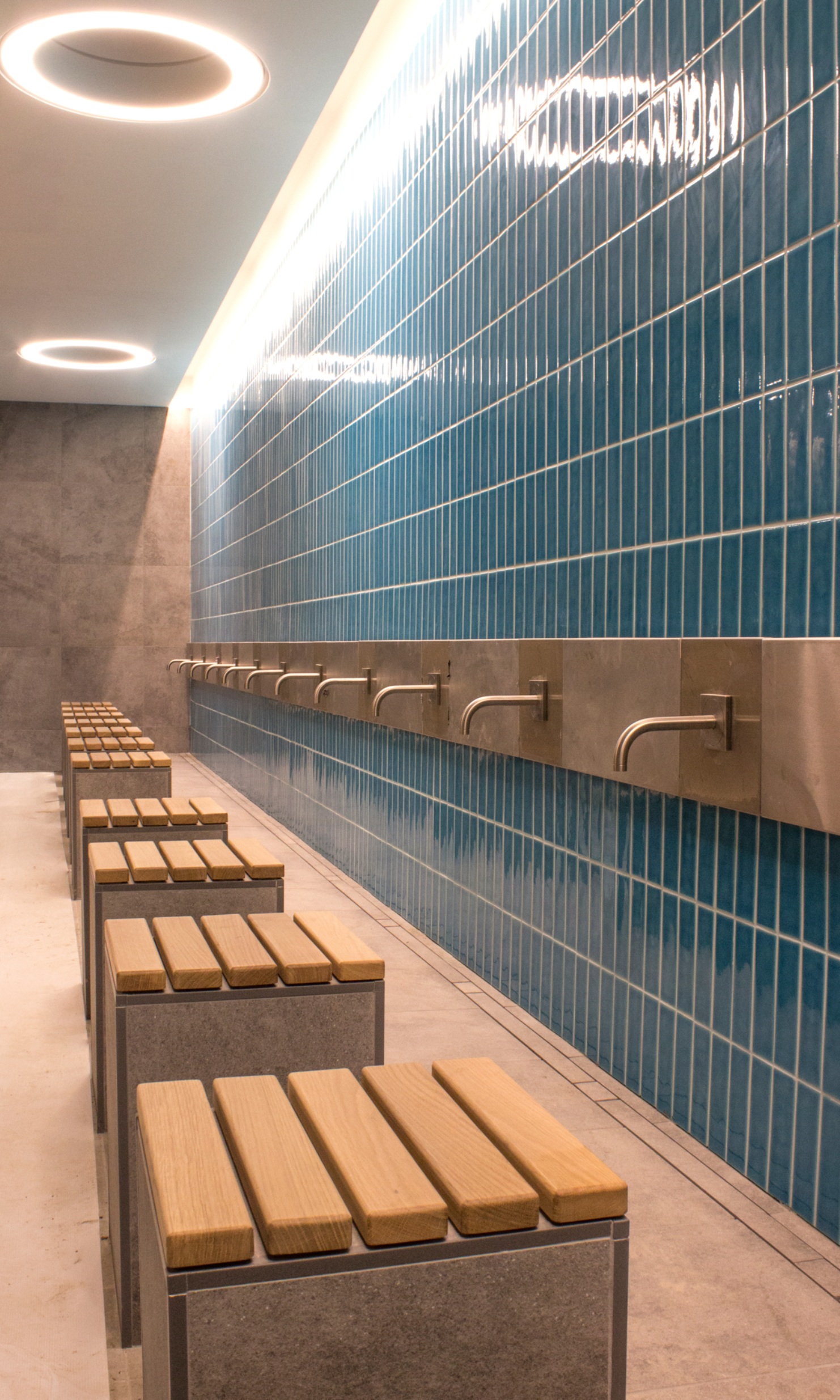 La prise de vue montre l’espace de lavage de la mosquée de Cambridge. Les tabourets sont en pierre et recouverts de planches de bois. Les robinets sont fixés sur un mur en carrelage bleu.