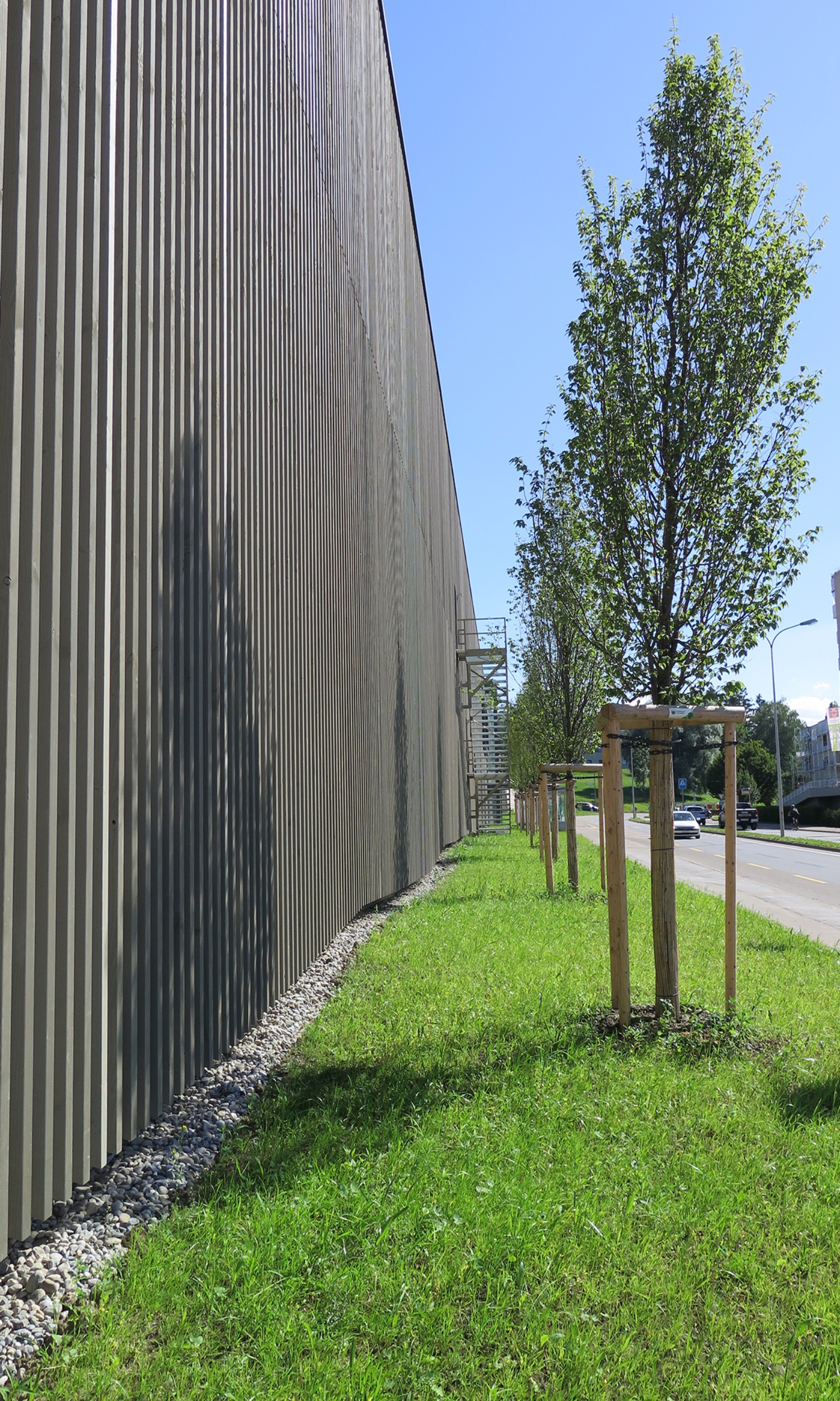 Seitliche Ansicht der Längsseite des Coop Super Center Uzwil mit dunkler Holzfassade, Rasen, jungen Bäumen und Strasse.