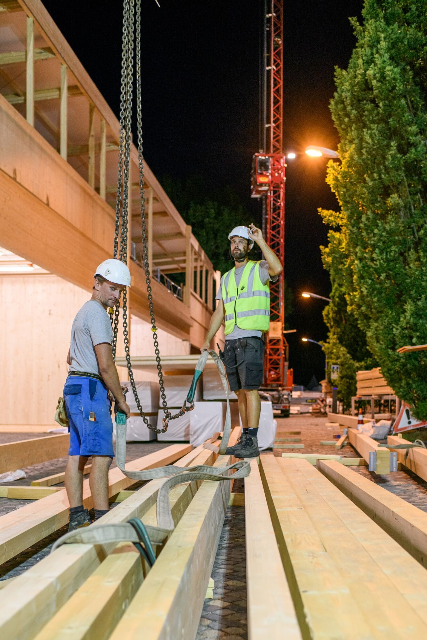 Während des Eventaufbaus befestigen und sichern zwei Arbeiter die Holzbalken an den Seilen von einem Kranen