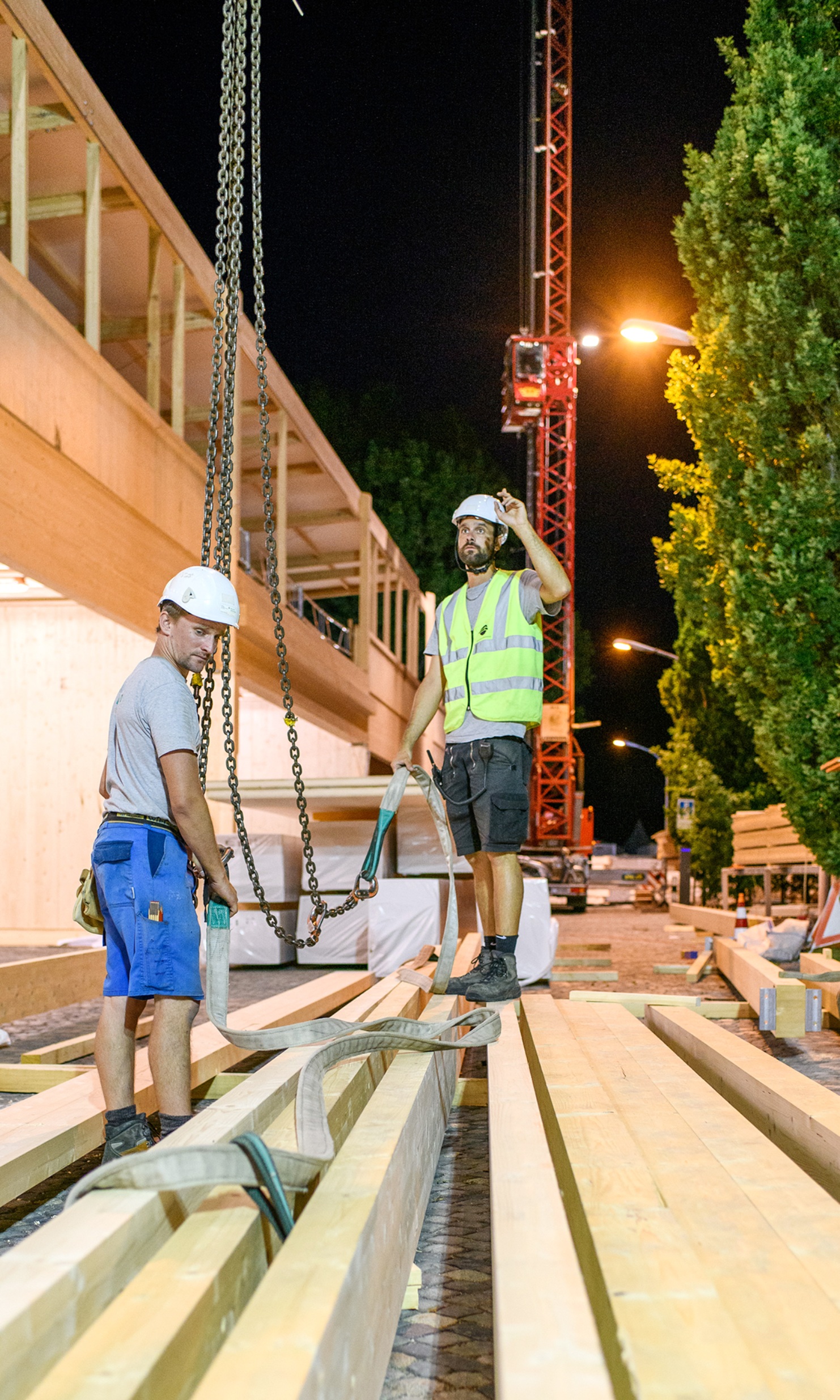 Two workers secure wooden beams with ropes hanging from a crane during the event construction