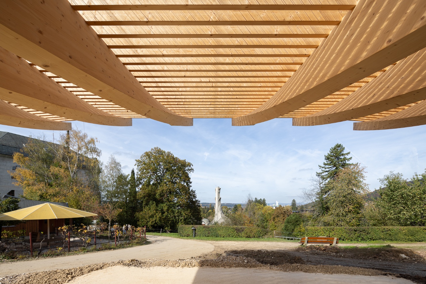 View from beneath the curved timber roof of the new Preparations House at the Goetheanum – in the background, the anthroposophically inspired site with its iconic buildings and autumnal park landscape.