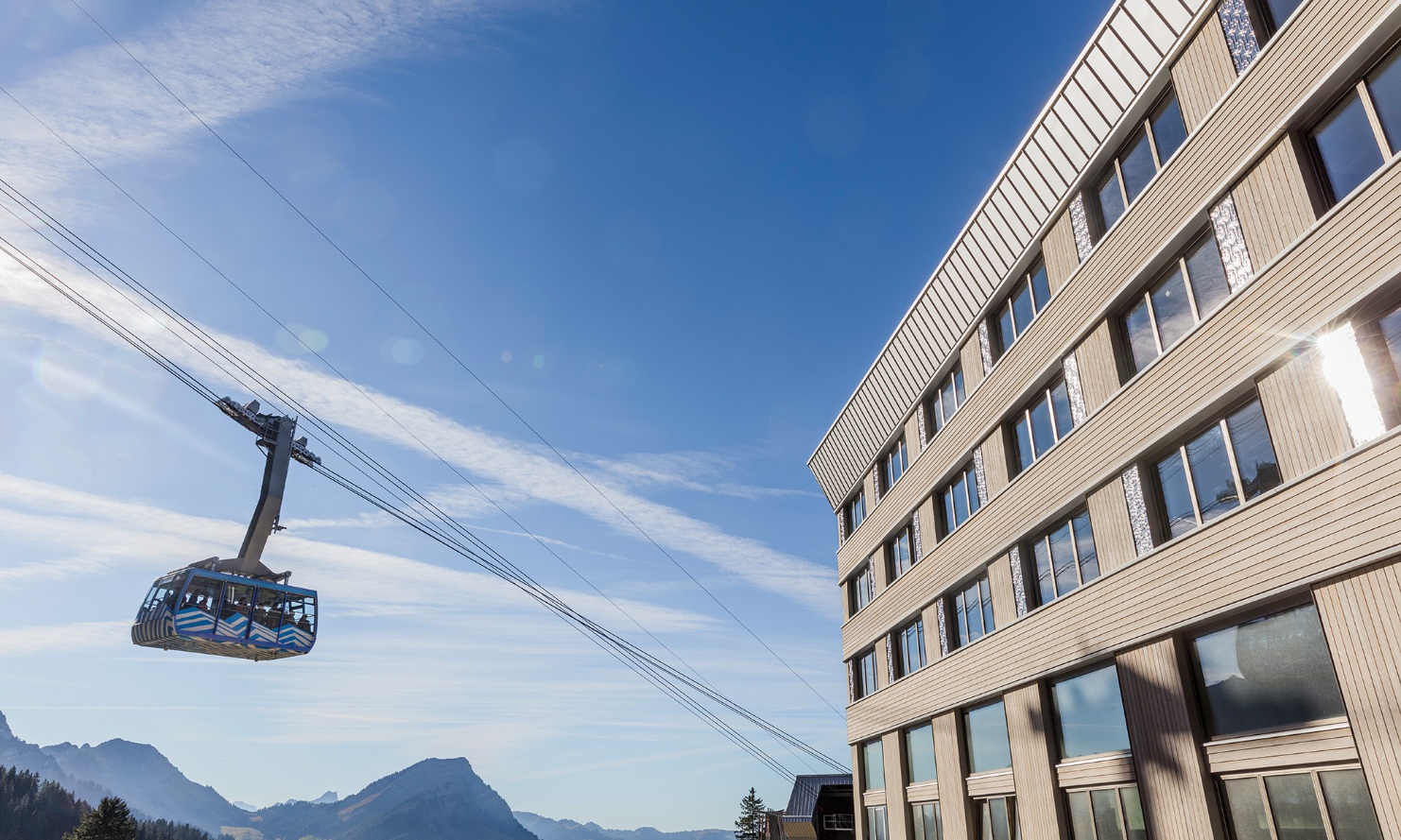 Aufnahme zeigt die Fassade vom Hotel Säntis sowie der von Besuchern genutzten Seilbahn. Im Hintergrund sieht man das Bergpanorama bei strahlend blauem Himmel.