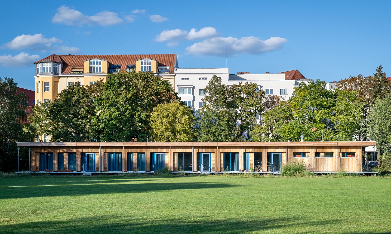 Visualisation of a single-storey, temporary school building in Berlin Schönefeld. The wooden facade stands out against the green meadow and blue sky.