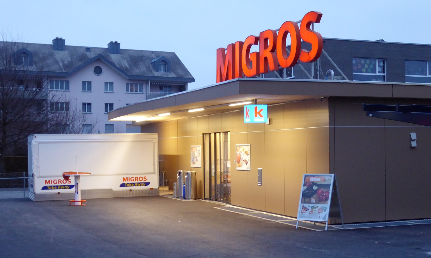 Ein eingeschossiger temporärer Holzbau in der Abenddämmerung mit der Migros-Leuchtschrift auf dem Dach.