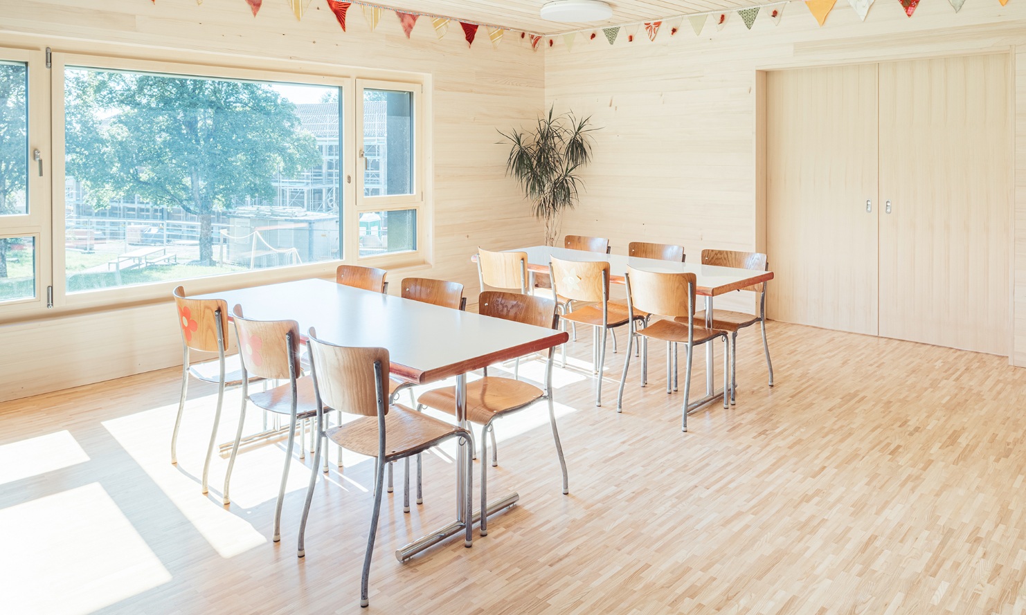 Salle à manger dans le nouveau bâtiment des structures d’accueil de jour. La grande baie vitrée latérale laisse entrer beaucoup de lumière naturelle dans la pièce au plancher, aux murs et au plafond en bois.