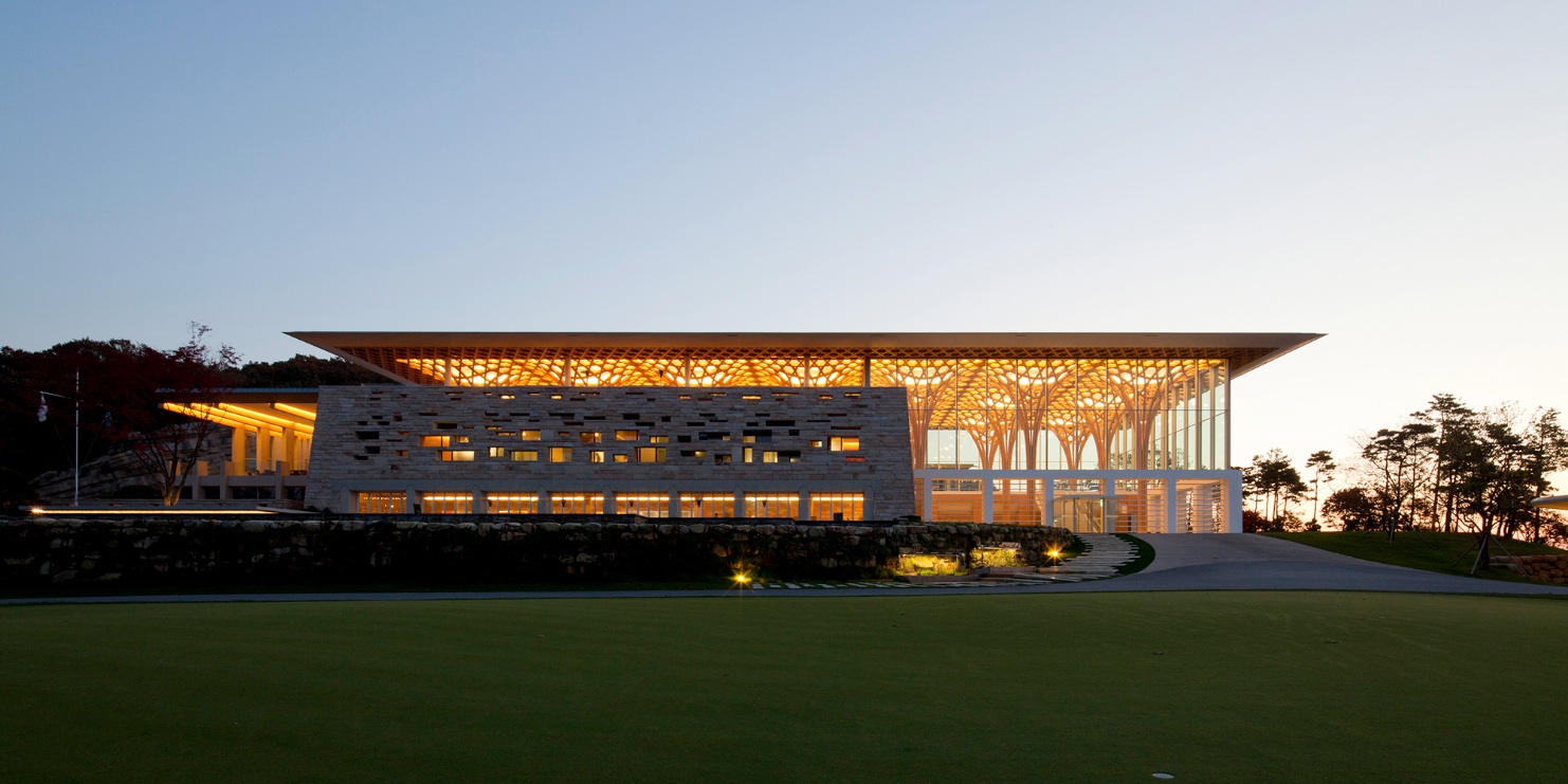 Vue du club-house illuminé au crépuscule. Les structures porteuses en bois, en forme d’arbres, sont visibles à travers les grandes façades vitrées.