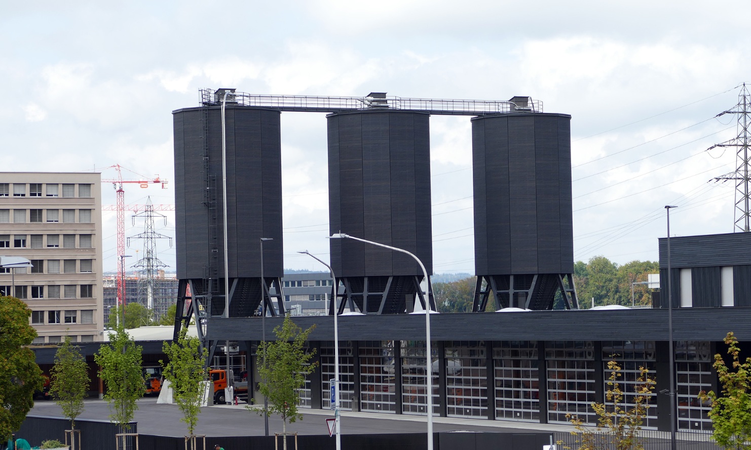 Three twelve-sided timber silos (E12), connected by a steel substructure and wooden roof platform, behind a fleet of trucks