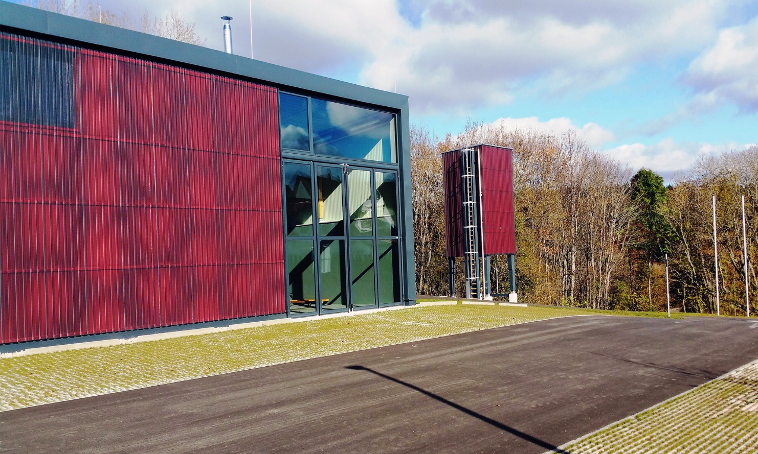 Moderner freistehender roter viereckiger Silo (E4) aus Holz neben neuem Gebäude mit roter Holzfassade und grossen grünen verglasten Toren