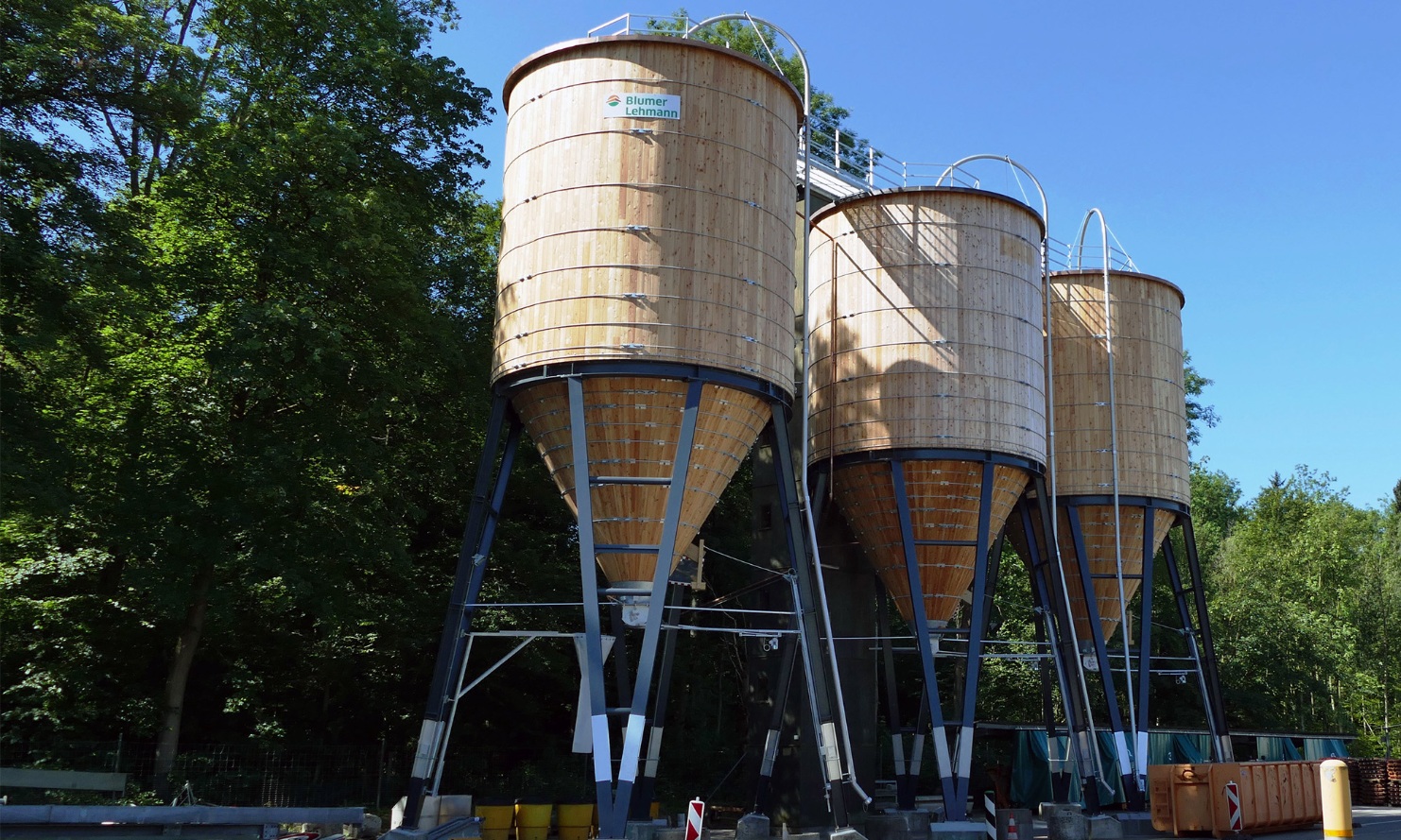 Three 200 m³ round timber silos with steel substructure, lined up in front of a group of trees