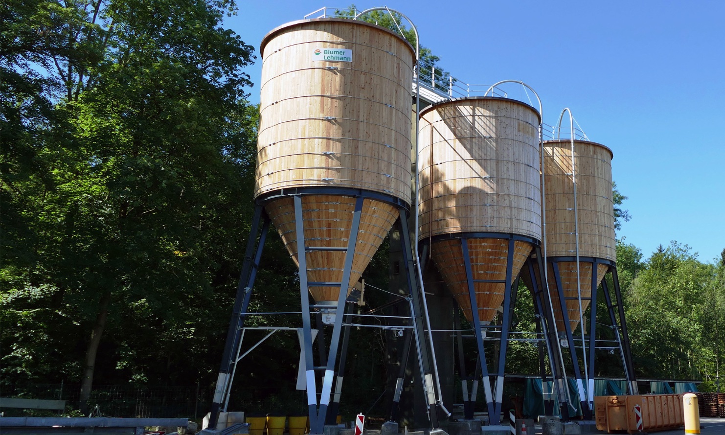 Drei runde 200 m³ Silos aus Holz mit Stahlunterbau nebeneinander vor einer Baumgruppe platziert 