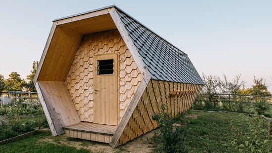 Wabenförmiges Bienenhaus mit Fassade aus sechseckigen Holzschindeln inmitten einer blühenden Wiese.
