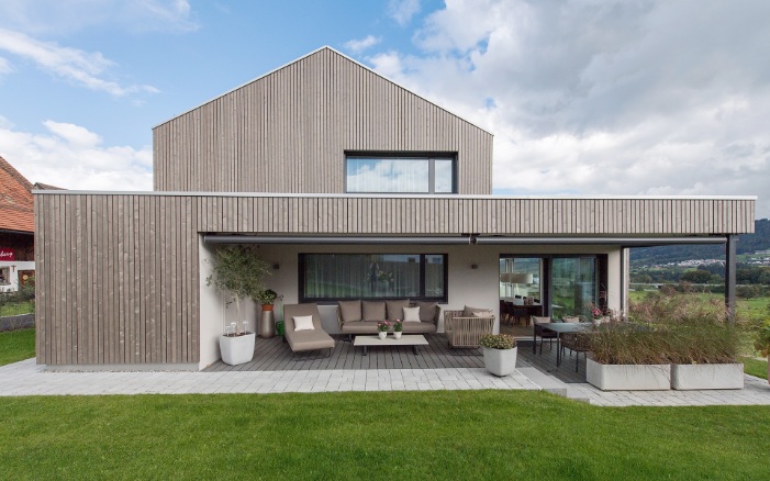 La prise de vue montre la maison individuelle avec vue sur le lac ainsi que sa terrasse couverte dans le jardin par beau temps