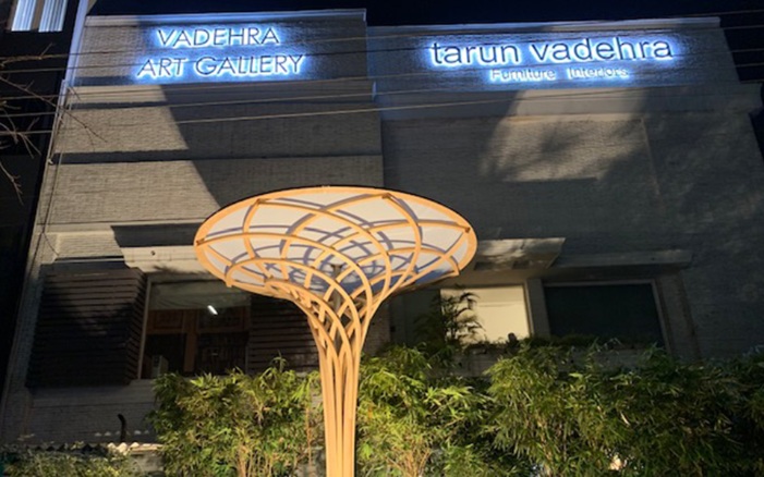 A free-formed wooden sculpture in the shape of a flower stands outside the Vadehra Art Gallery in Delhi. It is night and the free-form tree is illuminated.