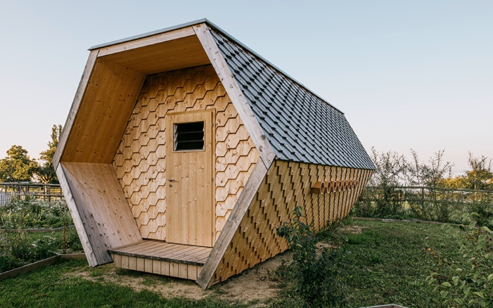 Wabenförmiges Bienenhaus mit Fassade aus sechseckigen Holzschindeln inmitten einer blühenden Wiese.