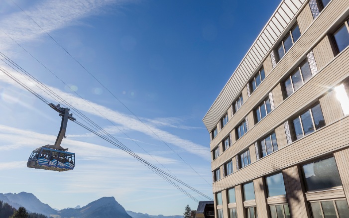 Aufnahme zeigt die Fassade vom Hotel Säntis sowie der von Besuchern genutzten Seilbahn. Im Hintergrund sieht man das Bergpanorama bei strahlend blauem Himmel.