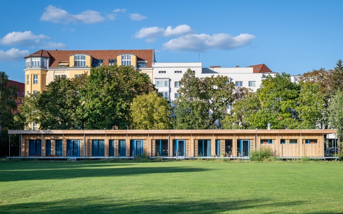Visualisation of a single-storey, temporary school building in Berlin Schönefeld. The wooden facade stands out against the green meadow and blue sky.