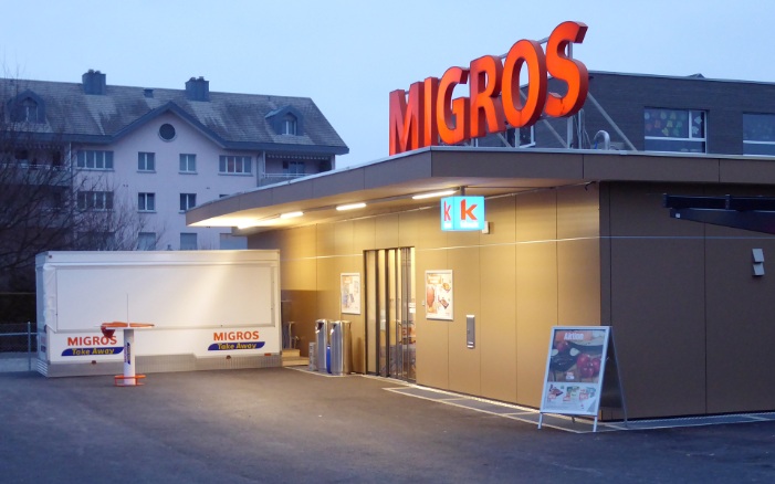 A single-storey temporary timber construction at dusk with the Migros neon sign on the roof.