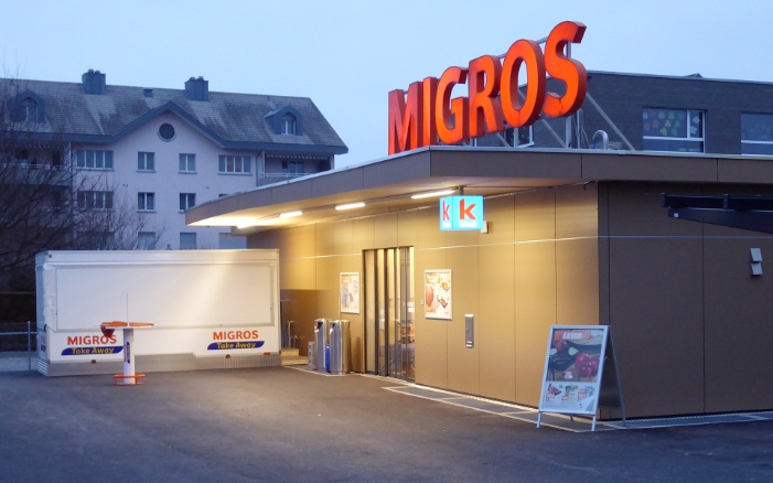 Ein eingeschossiger temporärer Holzbau in der Abenddämmerung mit der Migros-Leuchtschrift auf dem Dach.