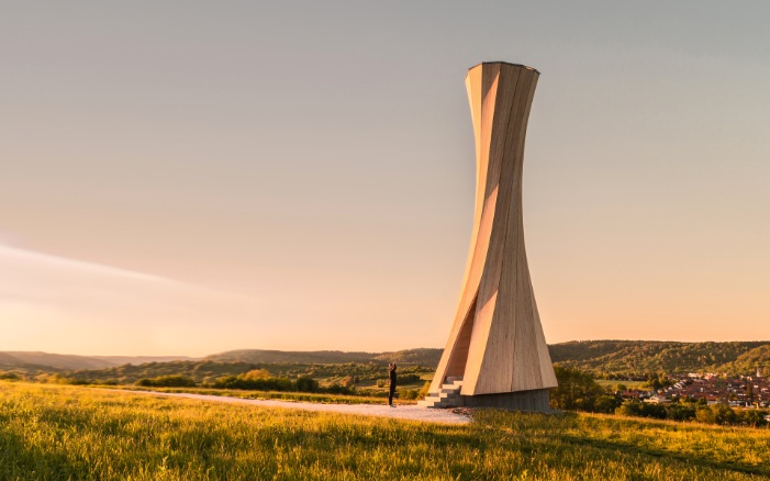 Gesamtansicht Urbach Tower bei Abendsonne. Die Grösse des Turms lässt sich gut erkennen, durch die Person, die neben dem Turm steht und sehr klein wirkt. Seine speziell geformte Holzstruktur fällt inmitten einer Hügellandschaft besonders auf.