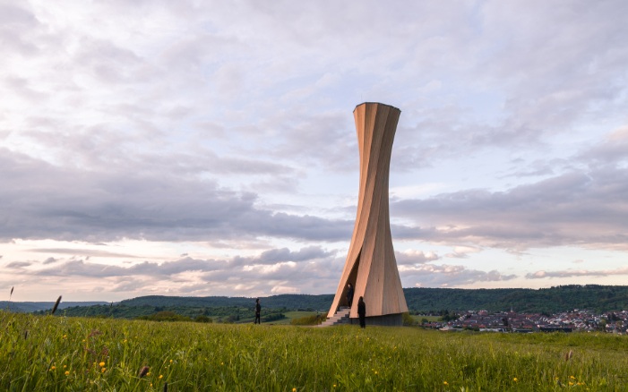 Gesamtansicht des Urbach Towers bei Morgendämmerung. Einige Leute besichtigen den Turm und seine speziell geformte Holzstruktur von innen und geniessen die weite Aussicht über die Hausdächer.