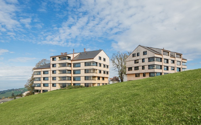 Two multi-storey timber residential buildings on a green meadow against a blue sky