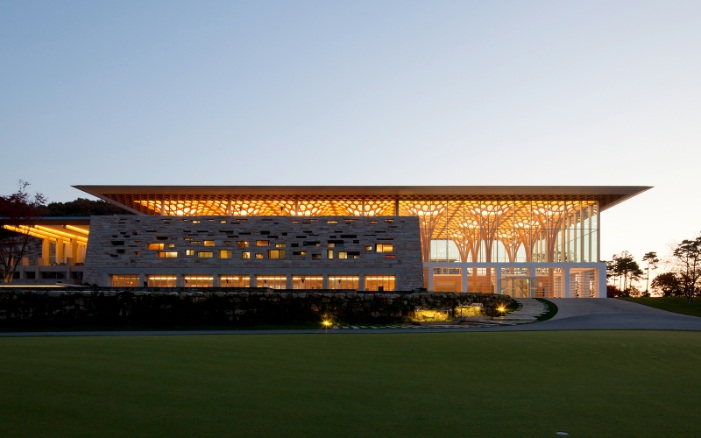 Vue du club-house illuminé au crépuscule. Les structures porteuses en bois, en forme d’arbres, sont visibles à travers les grandes façades vitrées.