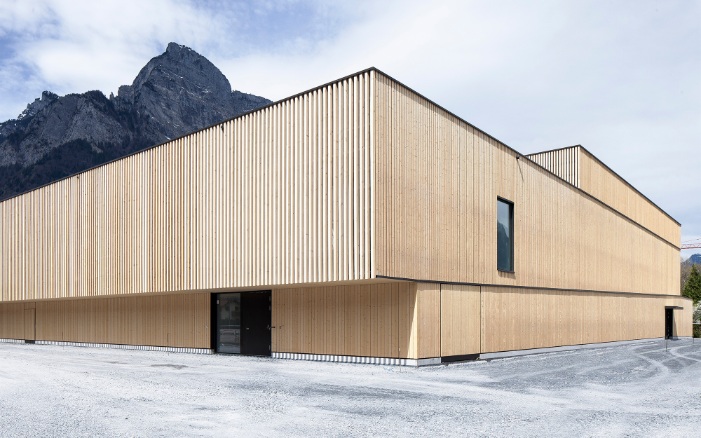 Frontansicht der Fassade einer Turnhalle mit blauem Himmel und Bergen im Hintergrund