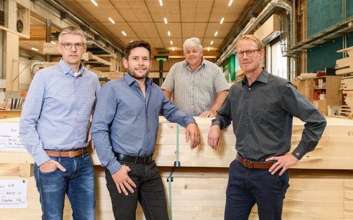 Markus Rutz, Martin Looser and Lukas Osterwalder stand in front of a stack of wood on the production site, with Richard Jussel behind. All four are looking at the camera in a friendly manner. 
