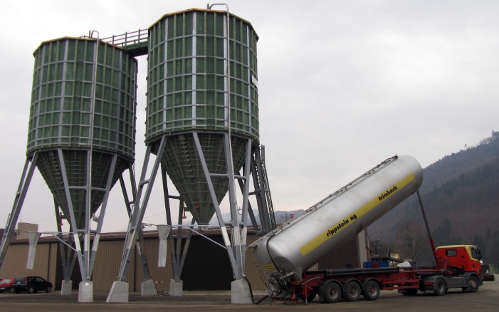 Deux silos dodécagonaux (E12) verts en bois de 400 m³, reliés par un support en acier et une plateforme de toit en bois, remplis par des camions-citernes