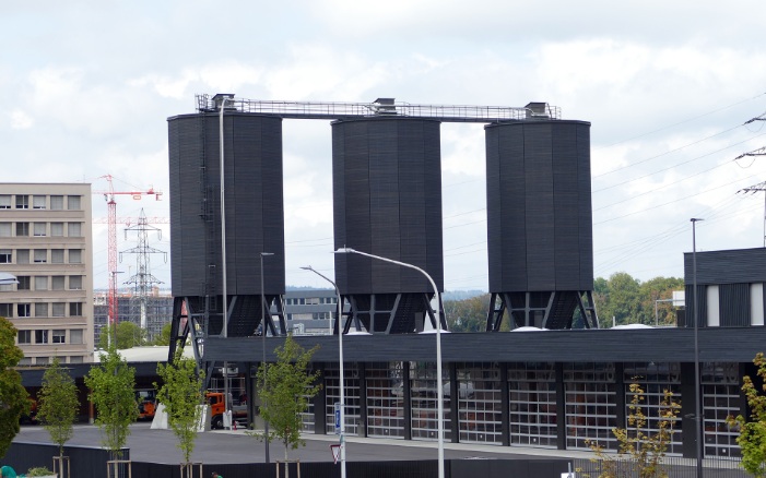 Three twelve-sided timber silos (E12), connected by a steel substructure and wooden roof platform, behind a fleet of trucks