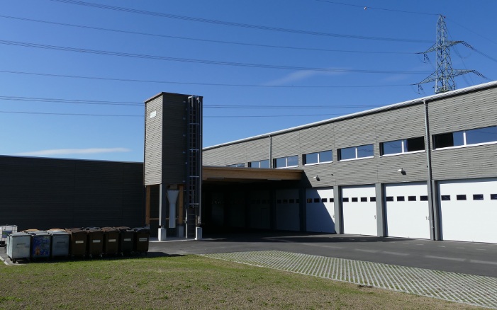 Grey, 40 m³ four-sided timber silo (E4) with steel substructure next to a large maintenance depot building with identical grey facade