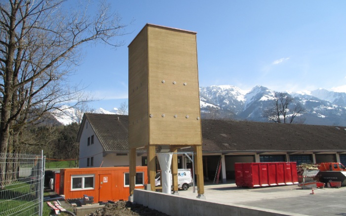 Silo carré (E4) en bois de 60 m³ avec support en bois et socle en béton devant l’ancien bâtiment de l’atelier et montagnes enneigées en arrière-plan
