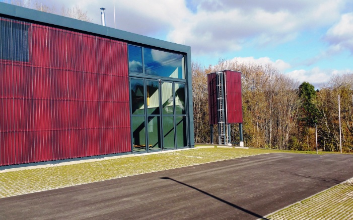 Moderner freistehender roter viereckiger Silo (E4) aus Holz neben neuem Gebäude mit roter Holzfassade und grossen grünen verglasten Toren