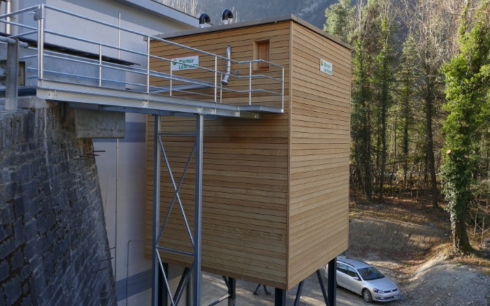 Silo modulaire en bois de 75 m³ avec support en acier gris et accès via une passerelle en acier, accolé à un bâtiment en bois existant