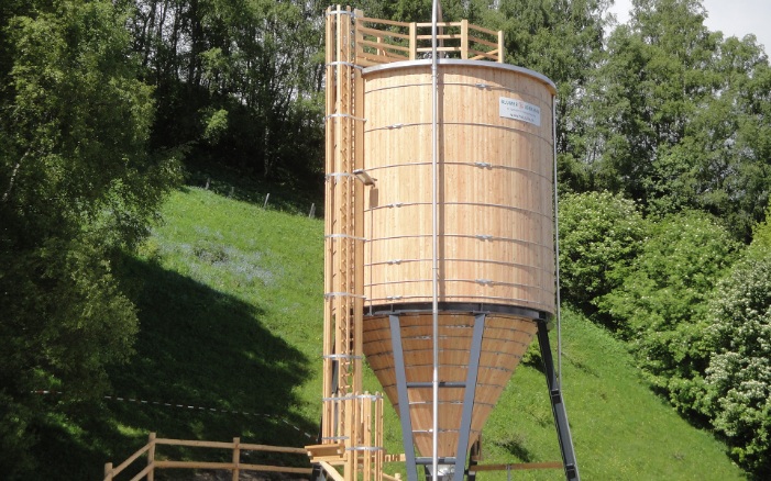 Runder 150 m³ Silo aus Holz auf schwarzem Stahlunterbau und Betonsockel vor grünem Hügel mit Bäumen platziert