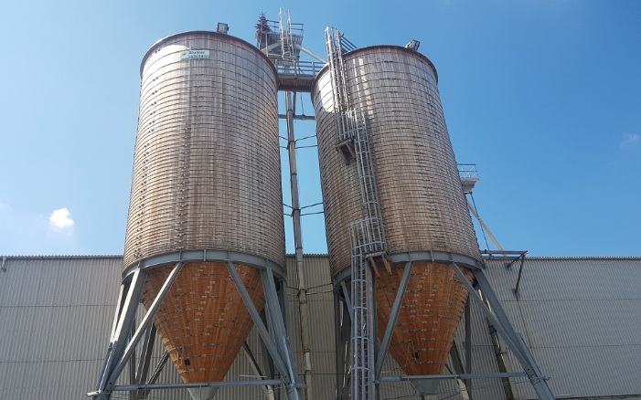 Two 600 m³ round timber silos with steel substructure and wooden ladder, connected by a timber roof platform, in front of an old industrial warehouse