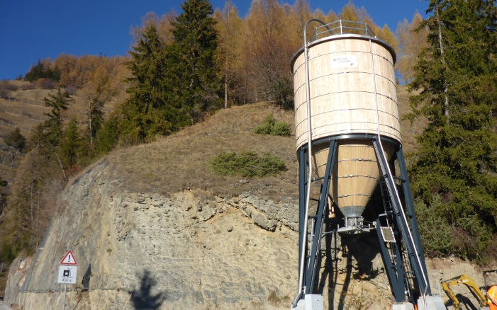 Silo rond en bois de 75 m³ posé sur un support en acier et un socle en béton au bord d’une route de col