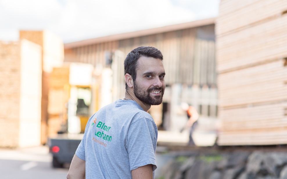 Ein junger Mitarbeitender mit grauem T-Shirt dunklem Haar und Bart schaut über die Schulter in die Kamera. Im Hintergrund sieht man Stapel mit geschnittenem Holz.