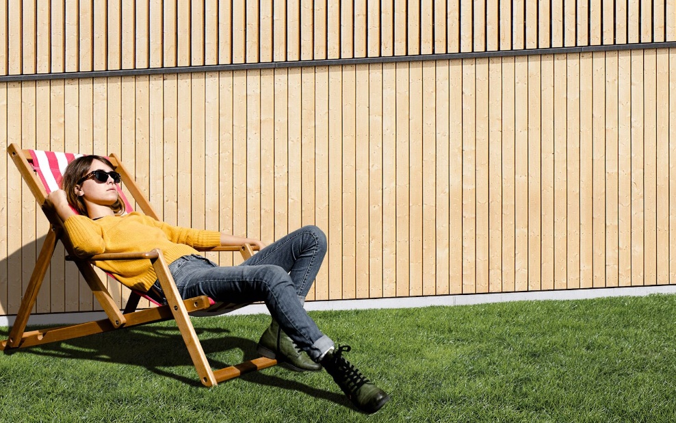 Commercial Holidays 2022 - Young woman lying in a deck chair in front of a wooden wall 