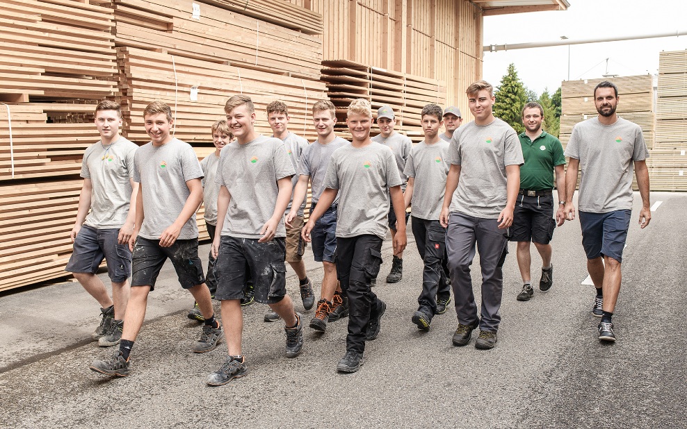 A group picture with eleven young people in training to become carpenters and two trainers walk in front of large stacks of wood and laugh