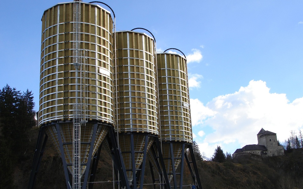 Trois silos dodécagonaux (E12) en bois avec échelle en acier et support en acier gris, posés sur un socle en béton avec un château historique en arrière-plan