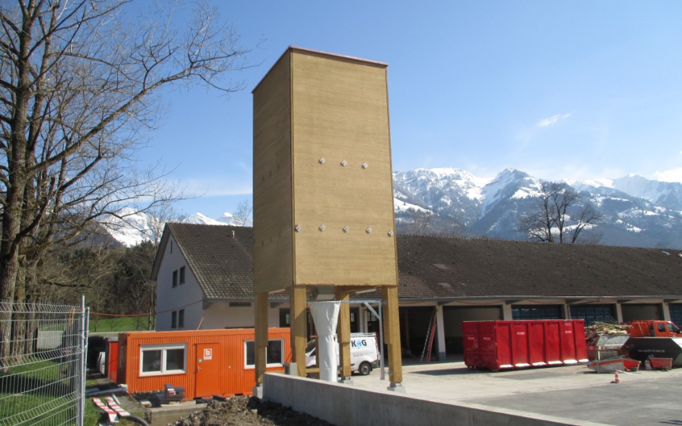 60 m³ four-sided timber silo (E4) with wooden substructure and concrete base, in front of a maintenance depot building with snow-capped mountains in the background