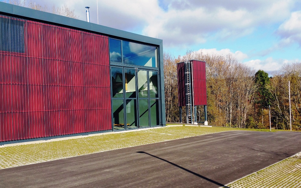 Moderner freistehender roter viereckiger Silo (E4) aus Holz neben neuem Gebäude mit roter Holzfassade und grossen grünen verglasten Toren